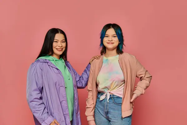 An Asian mother and her teenage daughter stand together in casual wear in front of a vibrant pink wall. — Stock Photo