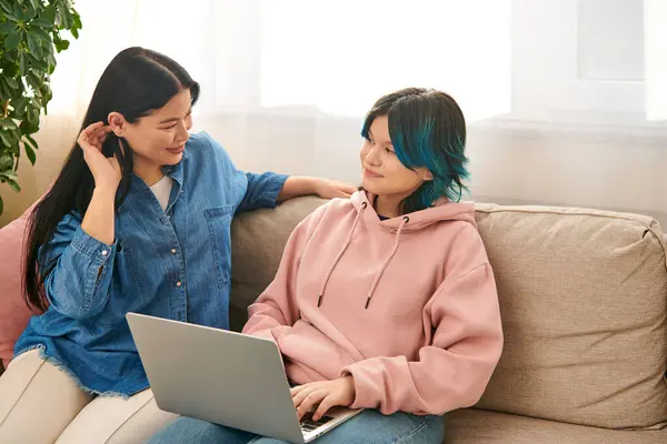 Una madre asiatica e sua figlia adolescente, in abbigliamento casual, si siedono su un divano, assorti mentre guardano entrambi uno schermo del computer portatile.. — Foto stock