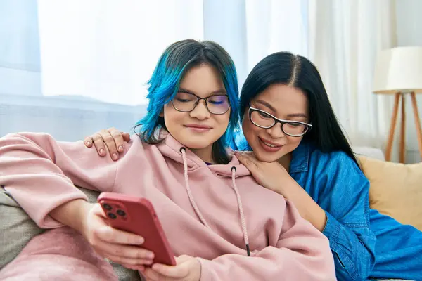 Two women, an Asian mother and her teenage daughter, sitting on a couch, engrossed in a cell phone. — Stock Photo
