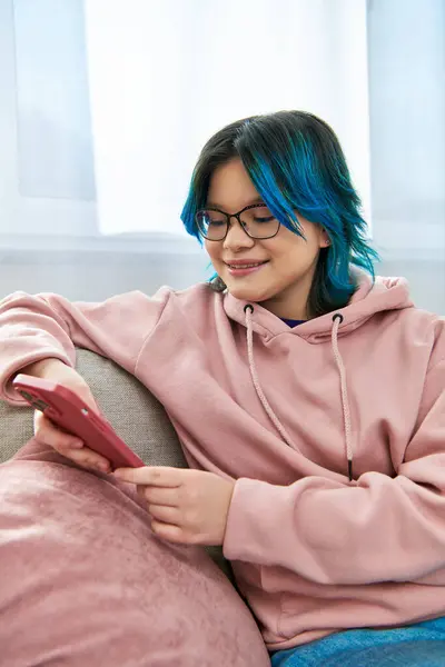 Une adolescente asiatique en tenue décontractée, assise sur un canapé avec téléphone, ayant des cheveux bleus frappants. — Photo de stock
