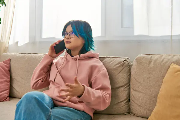 Une fille asiatique aux cheveux bleus s'assoit sur un canapé, parlant sur un téléphone portable ensemble. — Photo de stock