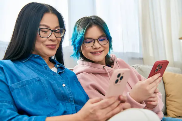 Asian mother and teenage daughter, in casual wear, sitting on a couch, focused on a cell phone screen in a cozy home setting. — Stock Photo