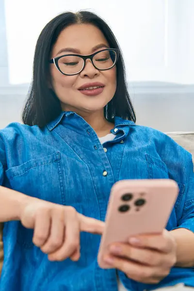 Une femme asiatique assise sur un canapé, engloutie dans un téléphone portable — Photo de stock