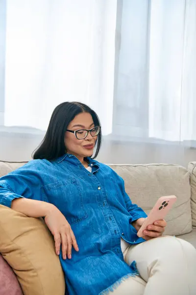 Uma mulher asiática de óculos sentada em um sofá, absorvida em celular — Fotografia de Stock