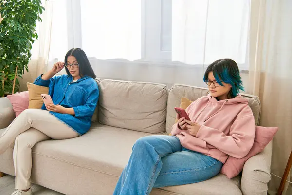 Asian mother and teenage daughter in casual wear sitting on a couch, engrossed in their phones, spending time together at home. — Stock Photo