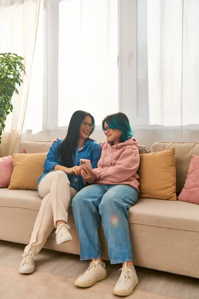 Una madre asiatica e sua figlia adolescente sedute giovialmente su un accogliente divano nel loro abbigliamento casual, legando e godendo di tempo di qualità insieme. — Foto stock