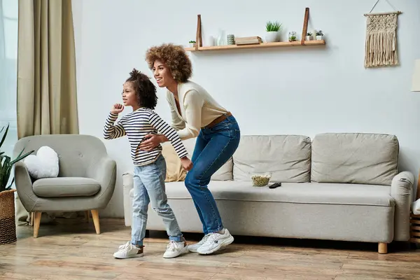 Una madre afroamericana feliz y su hija están de pie en un sofá en una acogedora sala de estar, compartiendo un momento especial juntos. - foto de stock