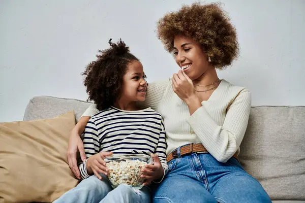 Bonne mère afro-américaine et sa fille assis sur un canapé, profiter d'une collation de pop-corn ensemble. — Photo de stock