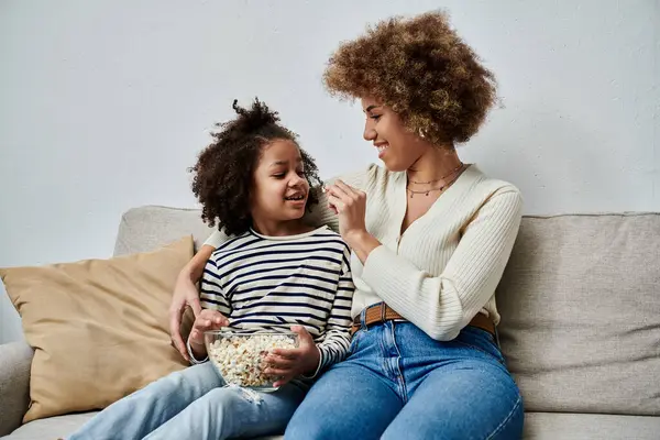 An African American mother and daughter happily sit on a couch, enjoying popcorn together. — Stock Photo