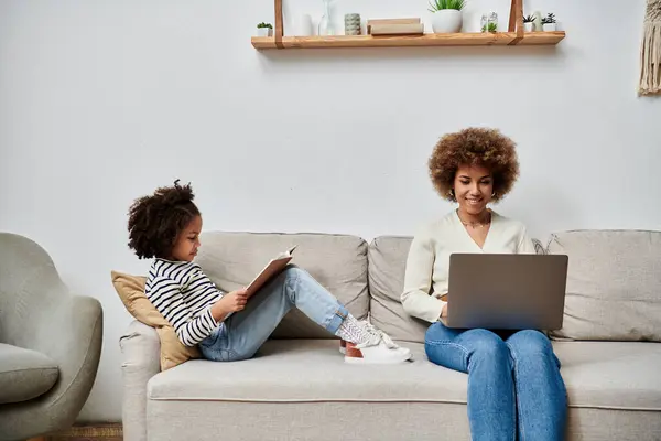 Une mère et une fille afro-américaine joyeusement assises sur un canapé, absorbées par l'utilisation d'un ordinateur portable ensemble. — Photo de stock