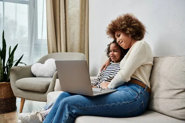 Une heureuse mère et fille afro-américaine assise sur un canapé, absorbée par l'utilisation d'un ordinateur portable ensemble. — Photo de stock