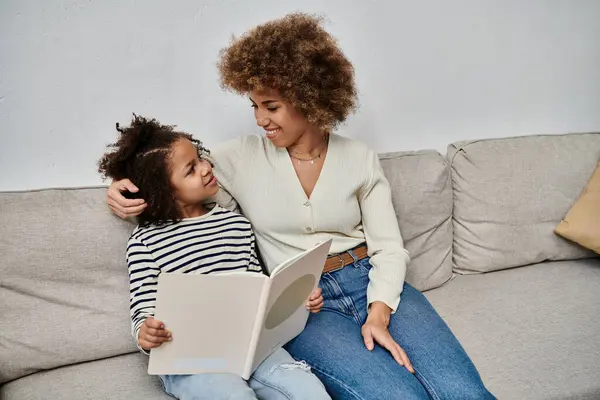 An African American mother and daughter sit together on a couch, engrossed in a book, sharing a moment of connection and togetherness. — Stock Photo