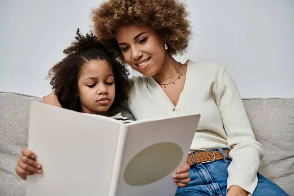 A happy African American mother and daughter sitting on a couch, immersed in a book, sharing a special moment together. — Stock Photo