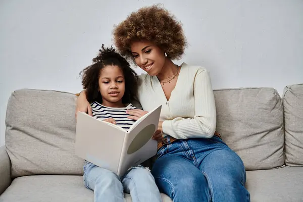 Uma alegre mãe e filha afro-americana imersa em um livro cativante enquanto sentada confortavelmente em um sofá. — Fotografia de Stock