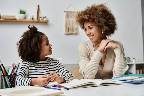 Une mère et une jeune fille afro-américaines sont assises à un bureau, engagées dans l'apprentissage et la liaison dans leur maison. — Photo de stock