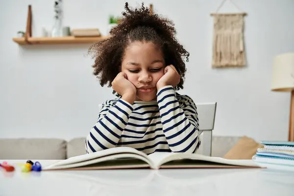 Bored African American girl engrossed in a book, sitting at a table — Stock Photo
