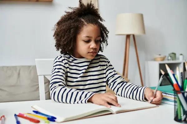 A young girl of African American descent sits at a table, engrossed in writing in a notebook — Stock Photo