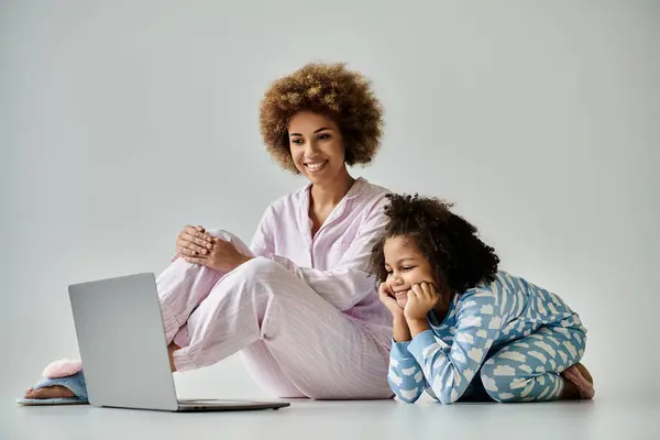 Una madre y una hija afroamericanas felices en pijama sentadas en el suelo usando un portátil. — Stock Photo