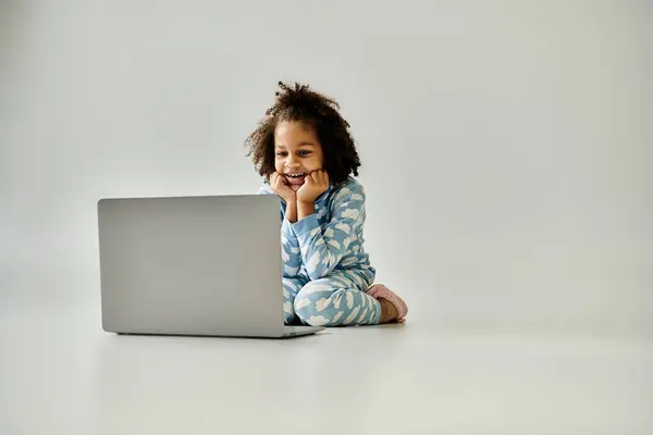 Une jeune afro-américaine en pyjama s'assoit sur le sol avec un ordinateur portable, explorant le monde numérique. — Photo de stock