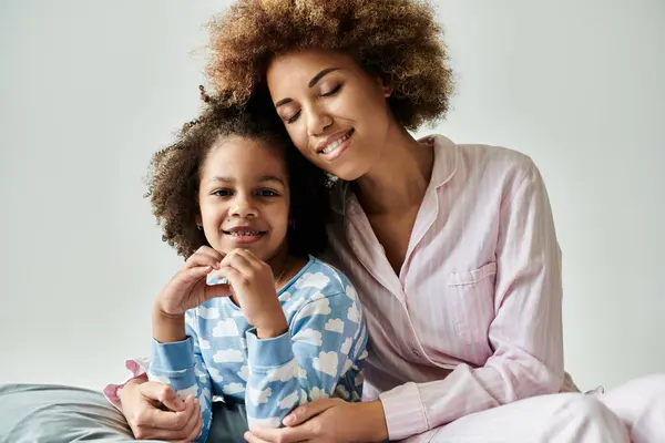 Mãe e filha afro-americana feliz em pijama, compartilhando um momento terno em uma cama aconchegante contra um fundo cinza suave. — Fotografia de Stock