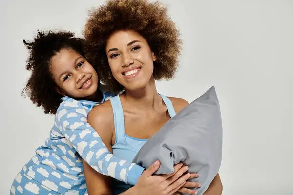Happy African American mother and daughter in pajamas sharing a loving hug with a pillow on a grey background. — Stock Photo