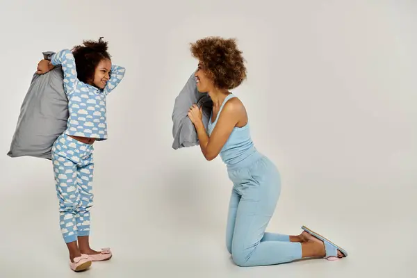 Happy African American mother and daughter in pajamas playfully enjoying a pillow fight on a light grey background. — Stock Photo