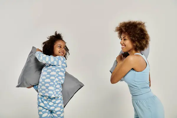 Happy African American mother and daughter in pajamas having a playful pillow fight on a white background. — Stock Photo