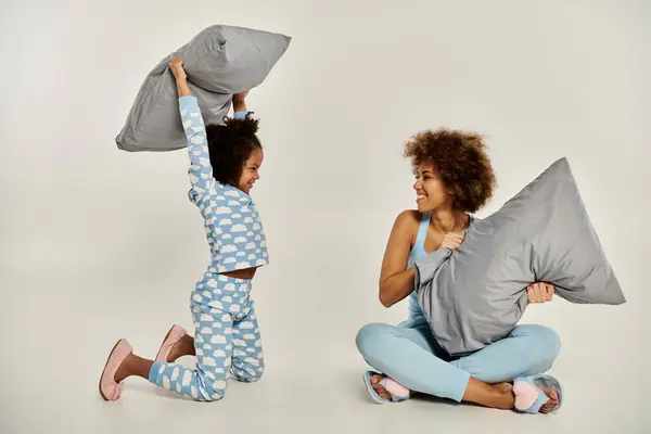 Uma feliz mãe e filha afro-americana de pijama se envolver em travesseiro brincalhão lutando em um fundo cinza. — Fotografia de Stock