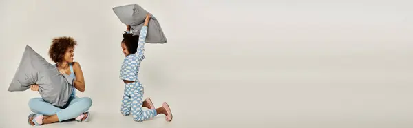 Joyful African American mother and daughter in pajamas playfully tossing pillows in a cozy moment on a grey background. — Stock Photo