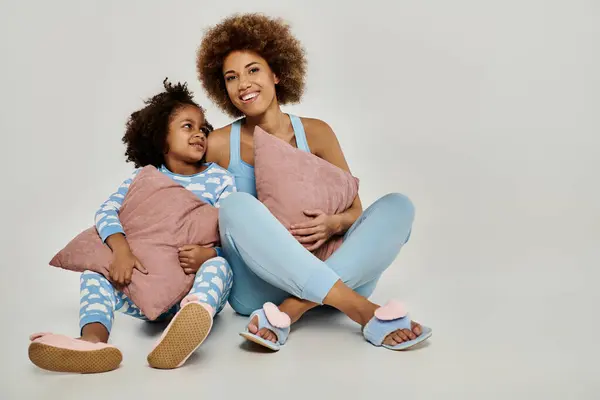 Bonne mère afro-américaine et sa fille assis sur le sol en pyjama, profiter d'un moment confortable ensemble sur un fond gris. — Photo de stock