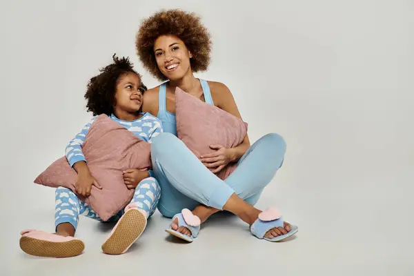 Happy African American mother and daughter sitting on the floor in pajamas, enjoying a cozy moment together on a grey background. — Stock Photo