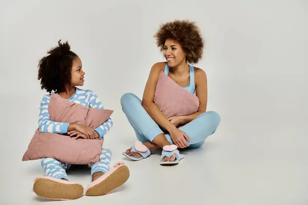 Duas mulheres afro-americanas felizes de pijama sentam-se no chão, segurando travesseiros, em um fundo cinza. — Fotografia de Stock