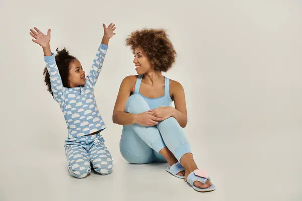 An ecstatic African American mother and daughter in pajamas sitting on the floor, arms raised to the sky in unison. — Stock Photo