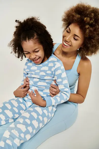 Mère et fille afro-américaine joyeuses portant un pyjama bleu assorti, profitant d'un moment ensemble sur un fond gris. — Photo de stock
