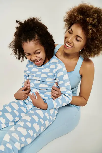 Cheerful African American mother and daughter wearing matching blue pajamas, enjoying a moment together against a grey backdrop. — Stock Photo