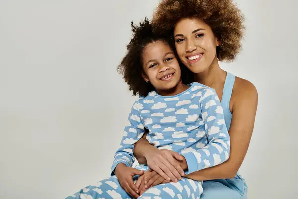 Joyful African American mother and daughter hugging tightly in cozy pajamas on a grey background. — Stock Photo