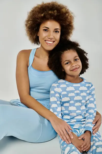 An African American mother and daughter smiling while posing in matching blue pajamas against a grey background. — Stock Photo