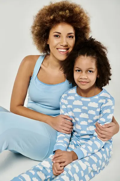 A joyful African American mother and daughter embrace, posing in matching blue pajamas against a soft grey backdrop. — Stock Photo