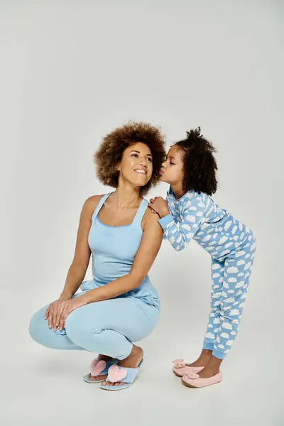 Un moment affectueux entre une joyeuse mère afro-américaine et sa fille, portant un pyjama bleu assorti, partageant un baiser. — Photo de stock