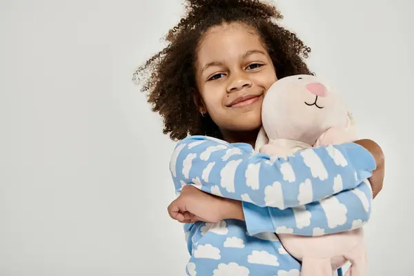 African American girl in pajamas cuddling a large stuffed animal on a grey background. — Stock Photo