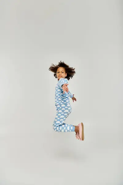 Une jeune fille énergique, vêtue de pyjama bleu, saute joyeusement dans les airs sur un fond gris. — Photo de stock