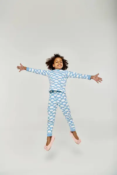 A happy African American girl joyfully jumping in a blue pajama set with her mother nearby on a grey background. — Stock Photo