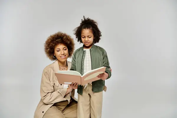 An African American mother reads a book to her little daughter, surrounded by love and joy in stylish clothing on a grey background. — Stock Photo