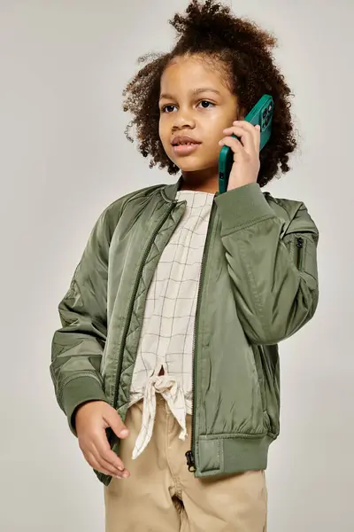 A young girl in a green bomber jacket talks on the phone, standing in stylish attire. — Stock Photo