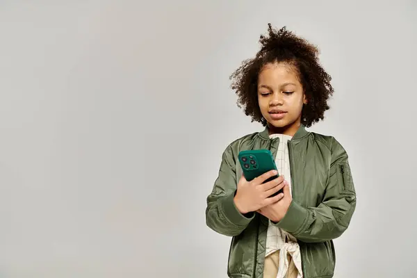 A stylishly dressed African American girl with curly hair holds a smartphone while standing against a grey background. — Stock Photo