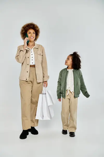 A curly African American mother and daughter, dressed stylishly, stand next to each other with shopping bags on a grey background. — Stock Photo