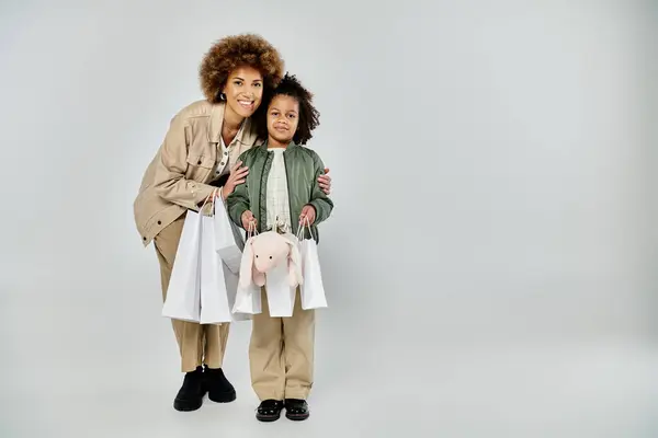 Curly African American mother and daughter in stylish attire, holding shopping bags on a grey background. — Stock Photo