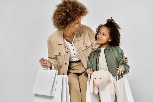 Um afro-americano encaracolado mãe e filha segurar sacos de compras, mostrando amor e alegria em um fundo cinza. — Fotografia de Stock