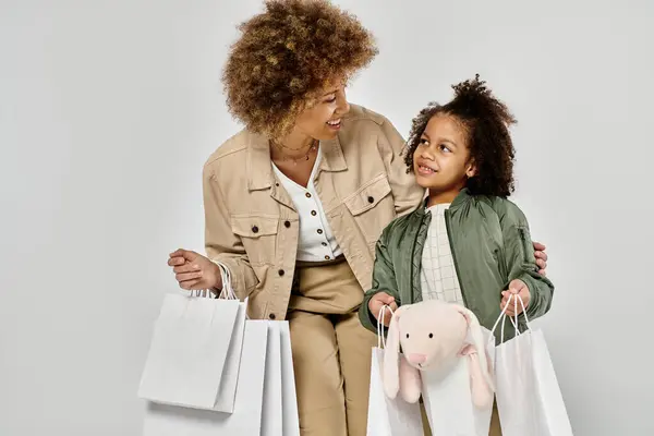 A curly African American mother and daughter hold shopping bags, showing love and joy on a grey background. — Stock Photo