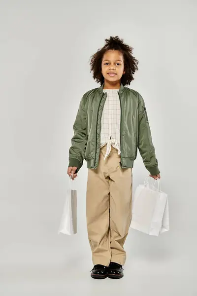 A stylish young girl with curly hair poses confidently while holding shopping bags on a gray background. — Stock Photo